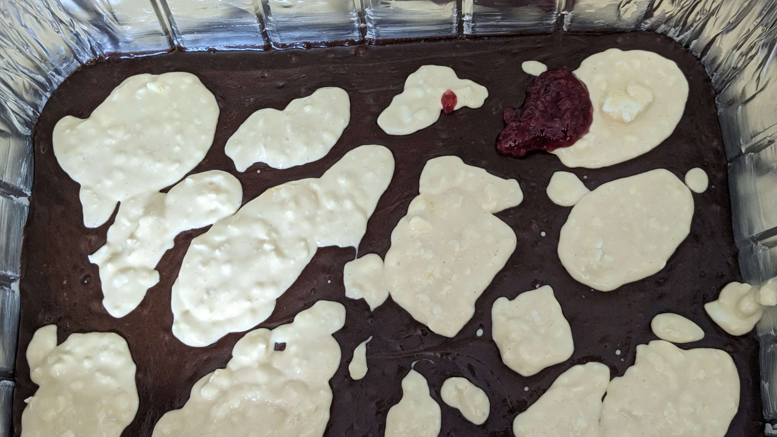 cream cheese mixture dolloped on brownie batter in an aluminum pan