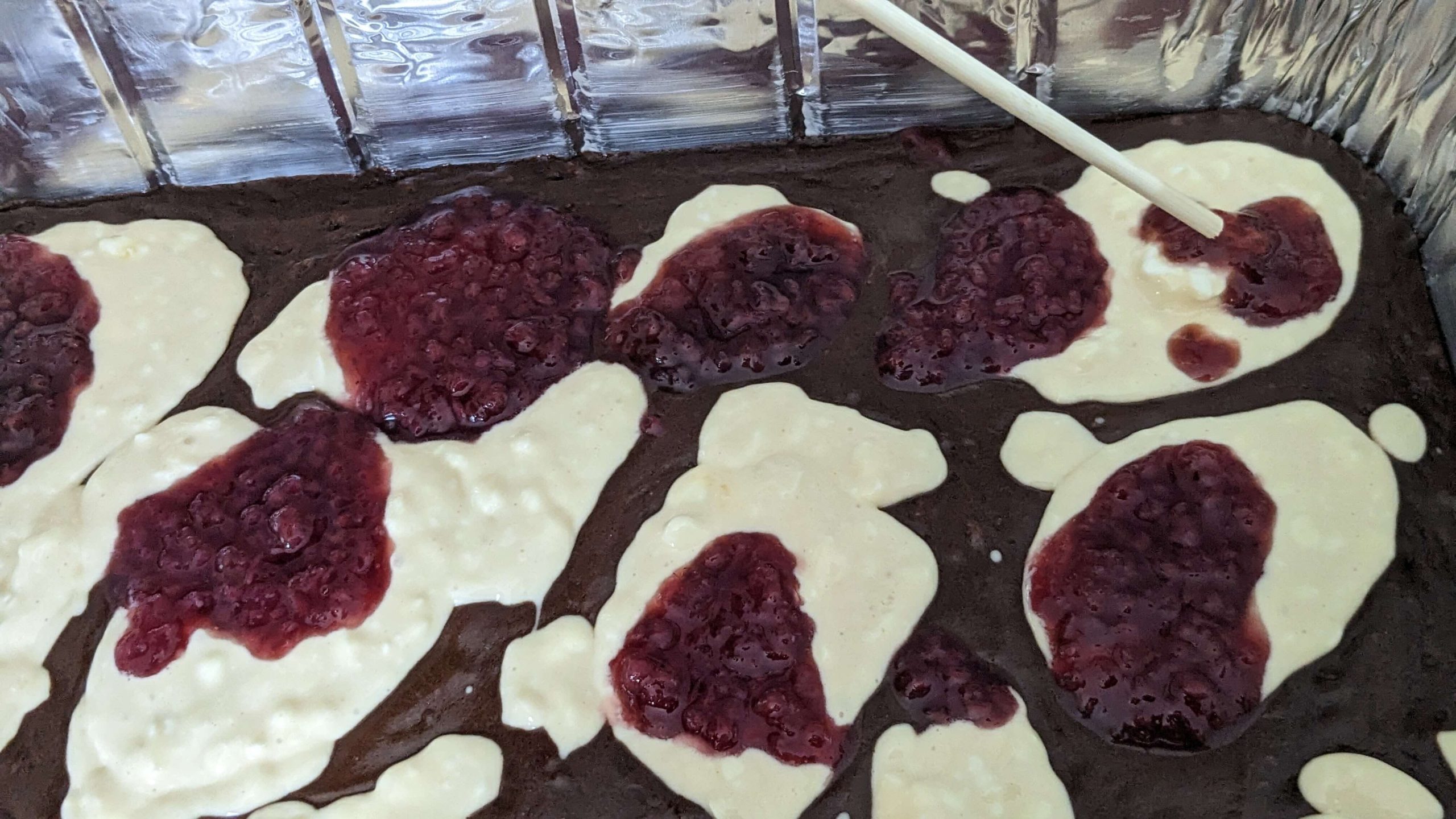 spoonfuls of raspberry jelly and cream cheese mixture dolloped on brownie batter in an aluminum pan