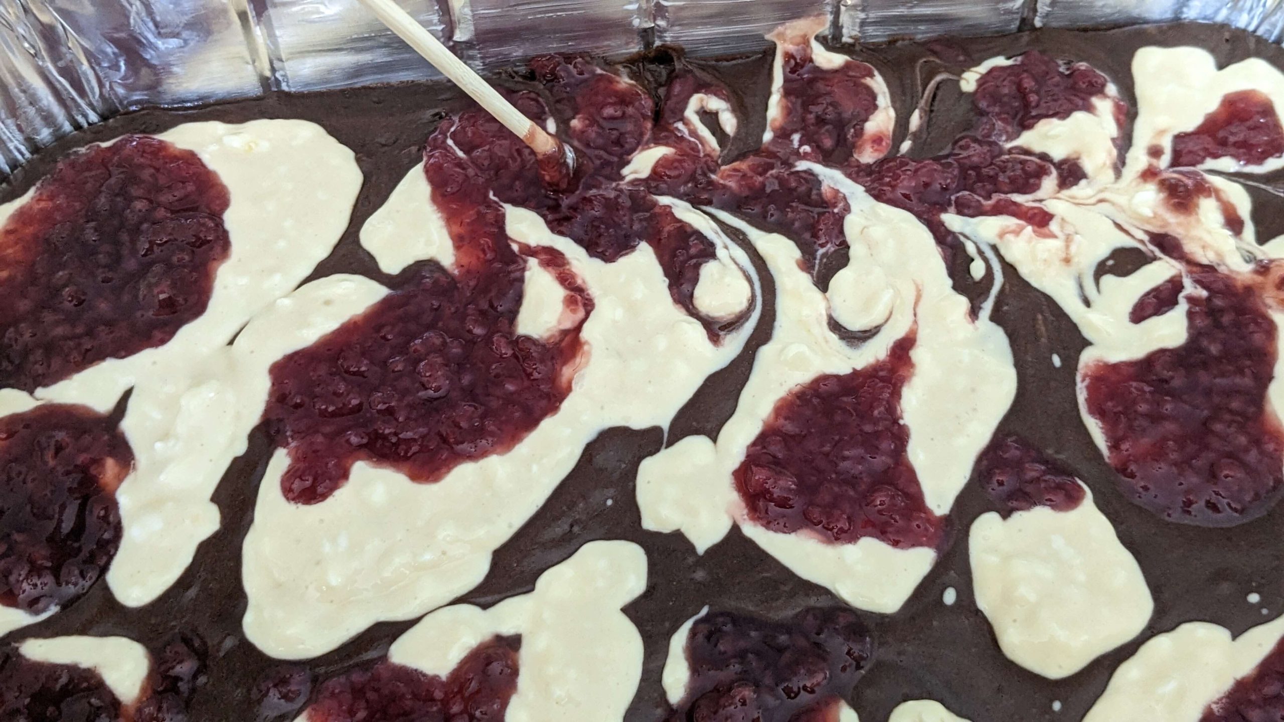 raspberry jelly and cream cheese mixture being swirled into brownie batter in an aluminum pan