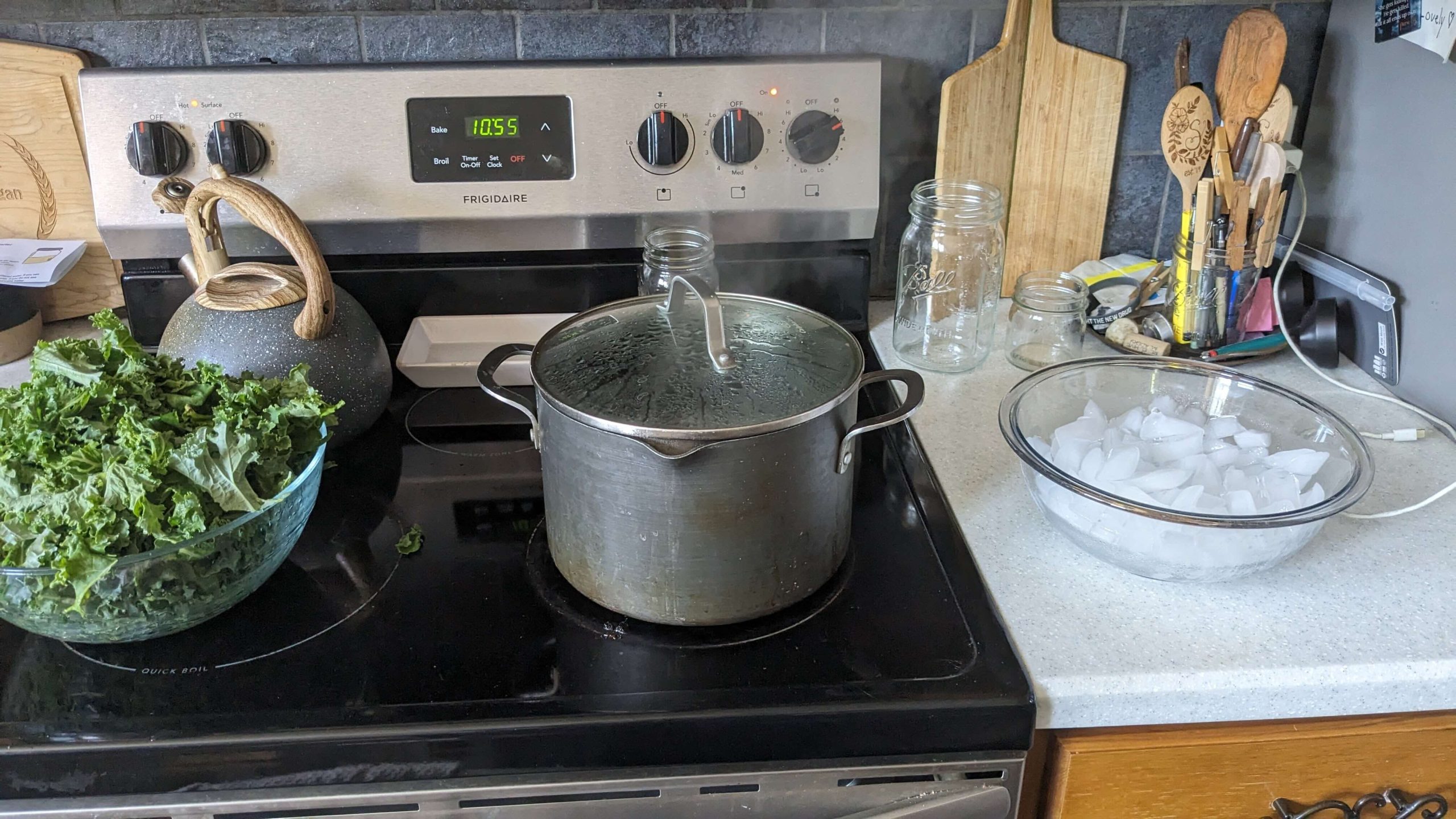 a bowl of raw kale next to a pot with a lid on it on the stove next to a bowl of ice