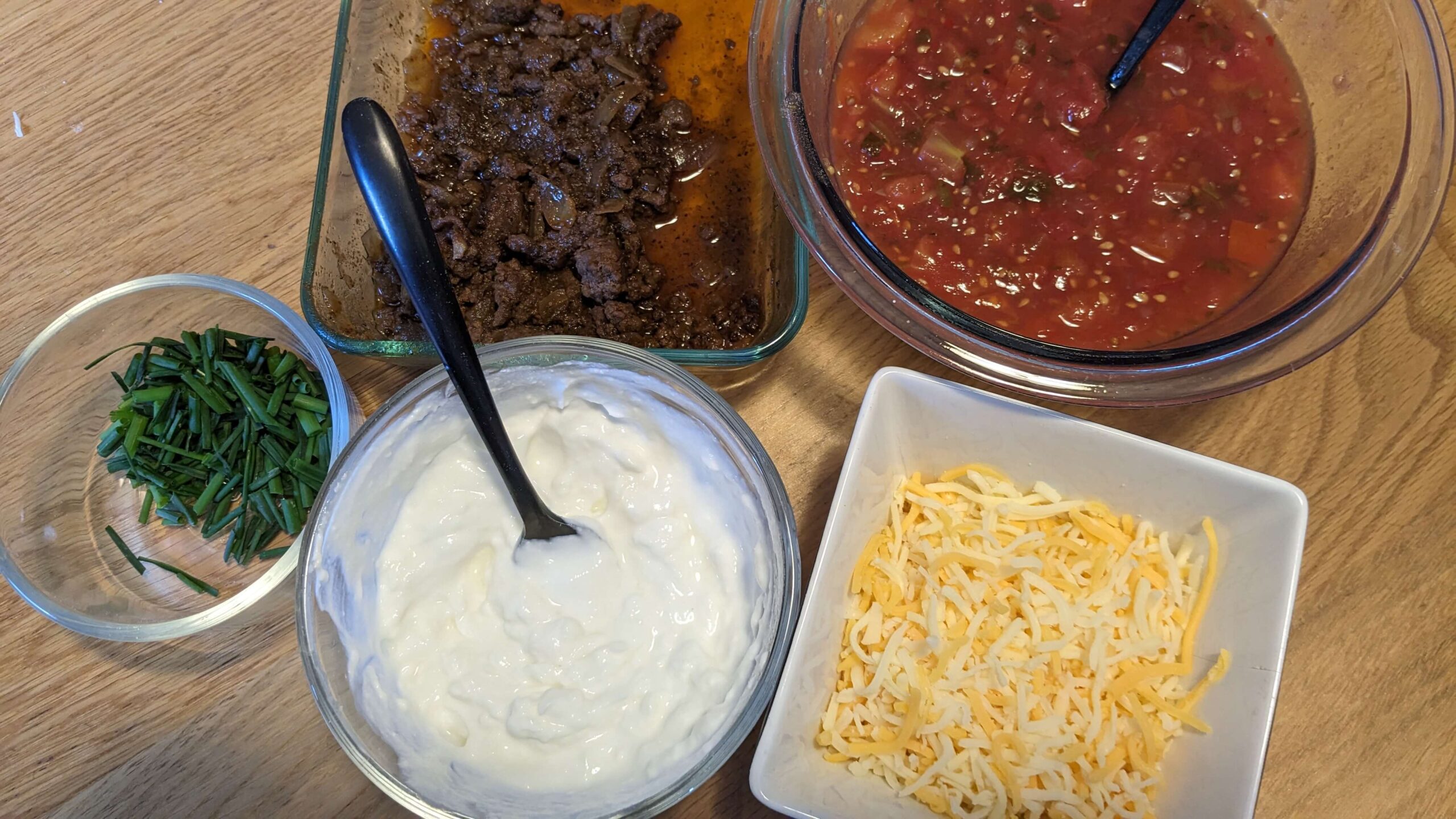 dishes of cooked taco meat, salsa, cut chives, cheese, and a bowl of sour cream with a black spoon in it 