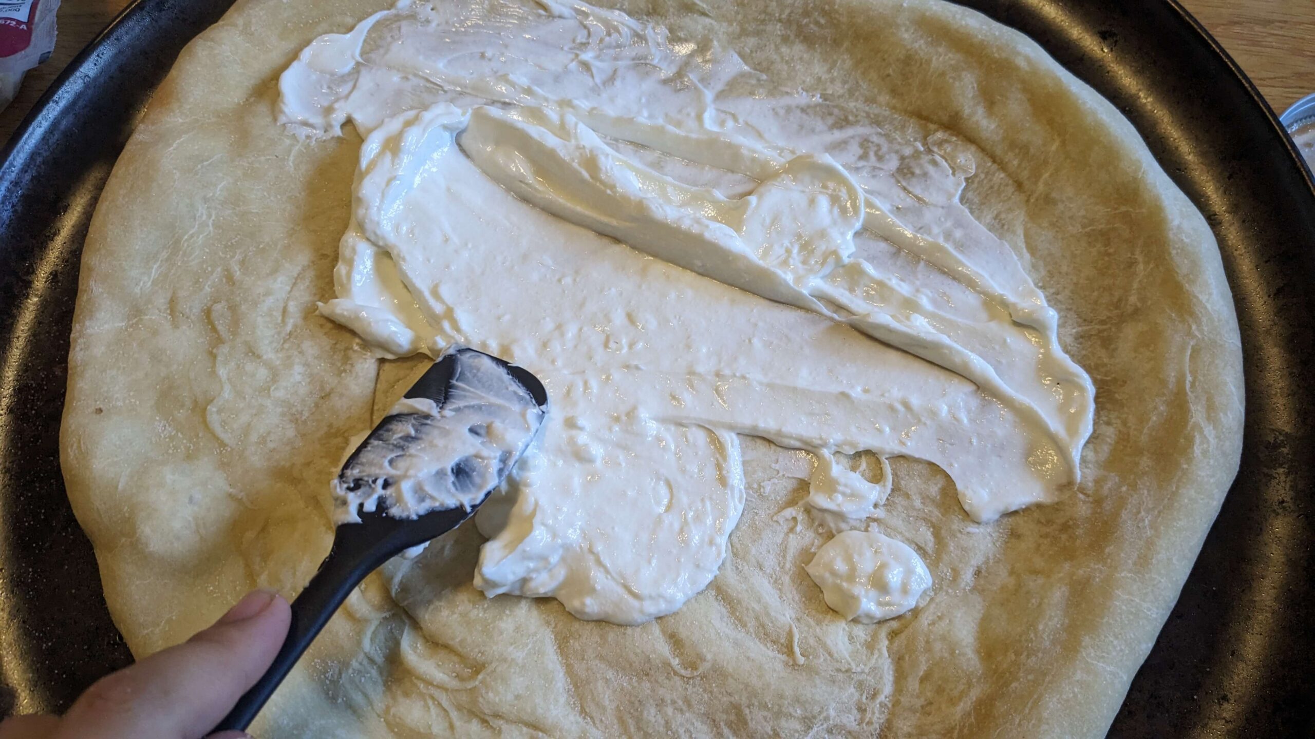 woman spreading a white mixture with a spatula on pizza crust 