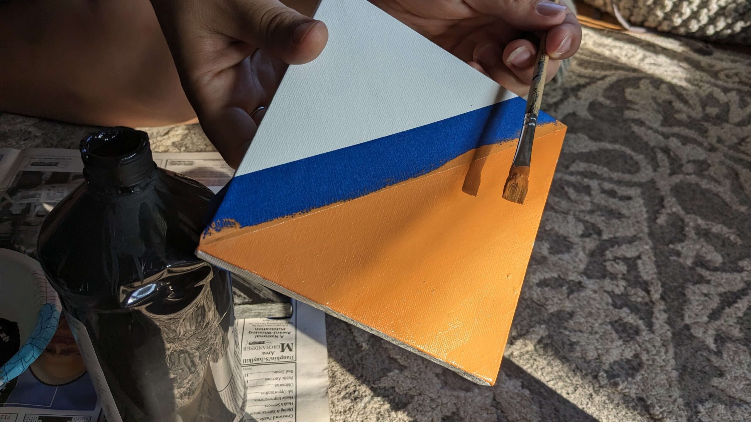 woman holding a square canvas with a diagonal blue painters tape strip and one half painted orange and holding a paintbrush