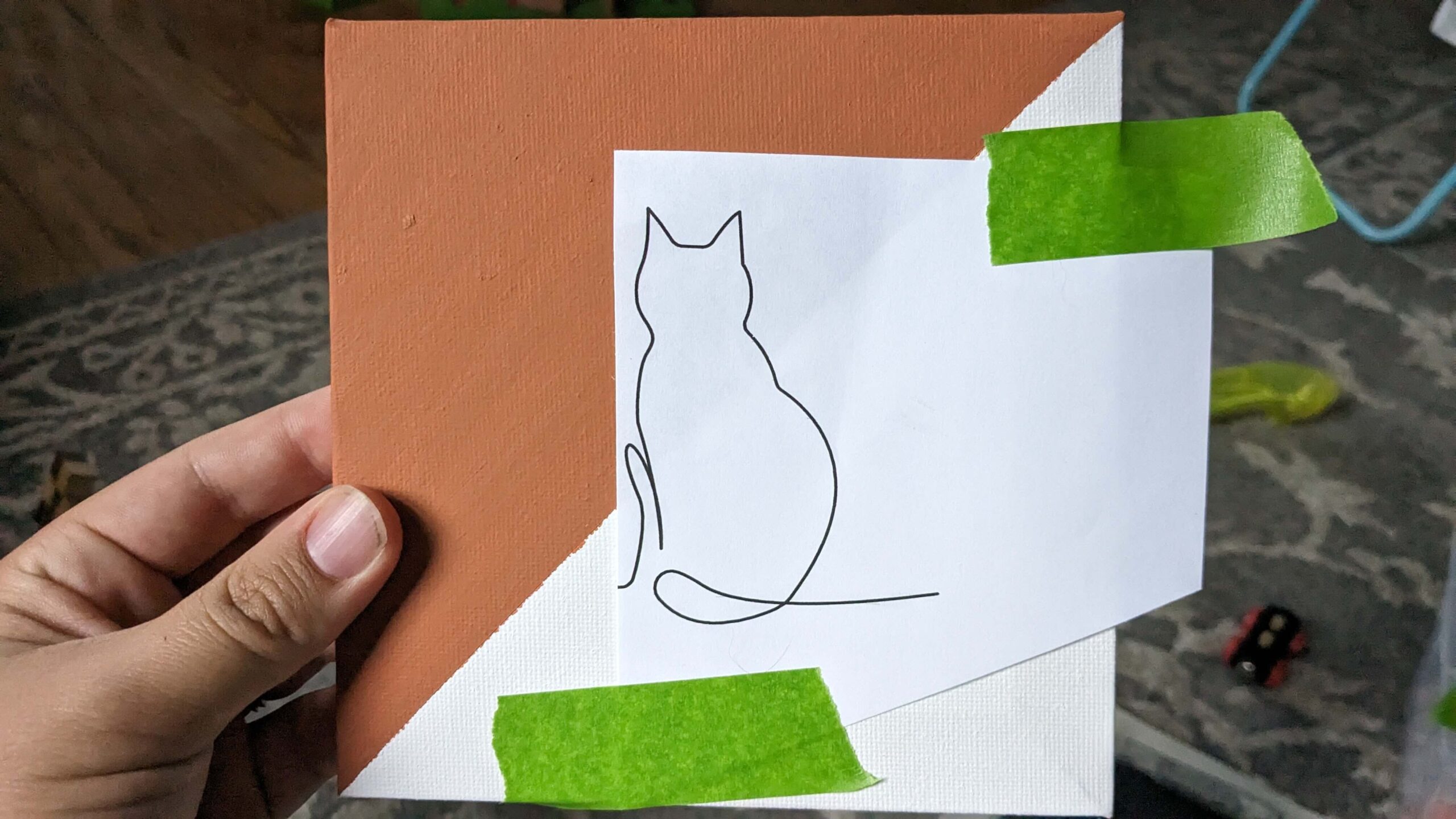 woman holding an orange and white square canvas with a cat line drawing taped to the center with green painters tape