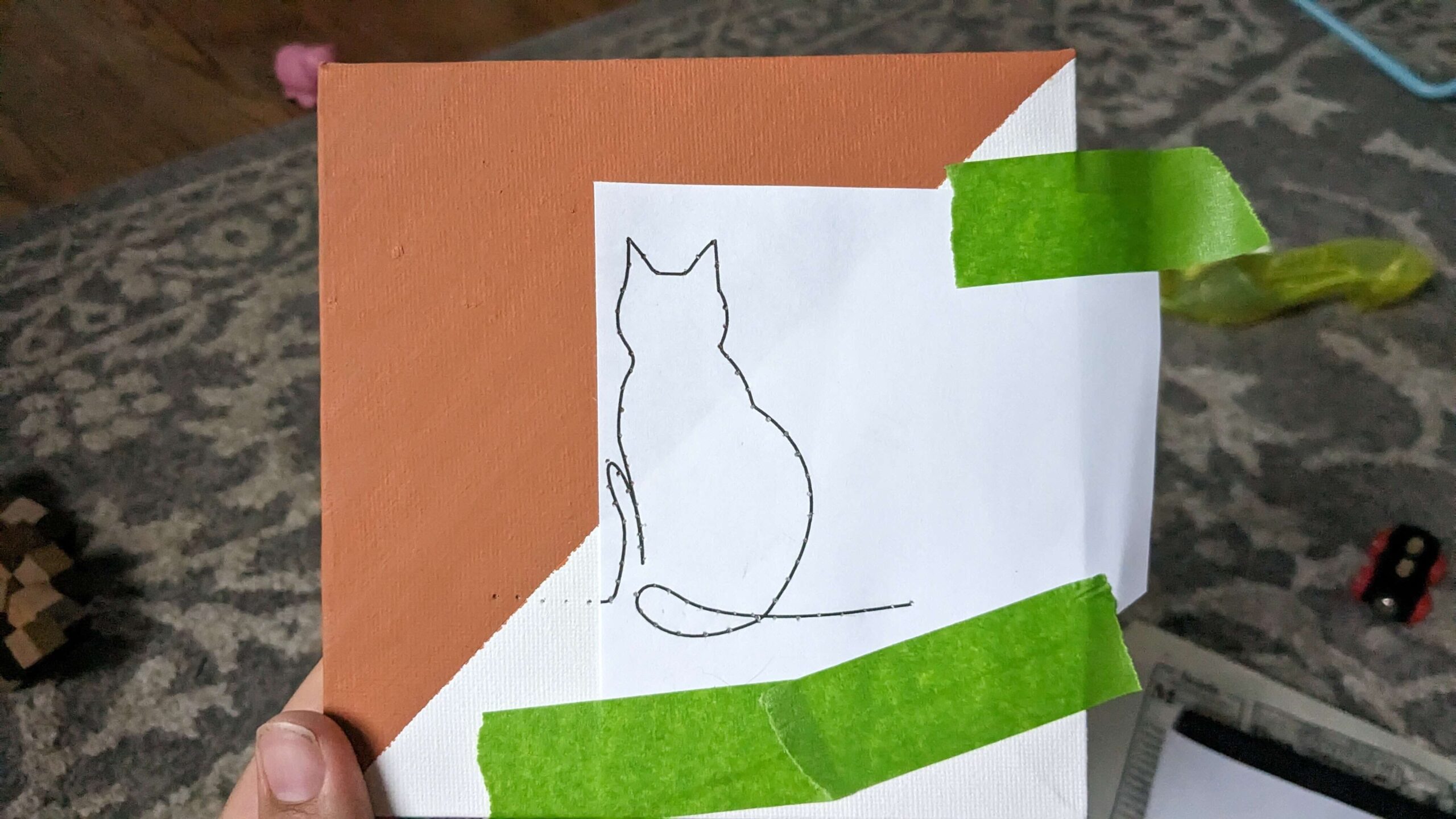 Woman holding an orange and white square canvas with a cat line art picture taped to it with green painters tape with small holes