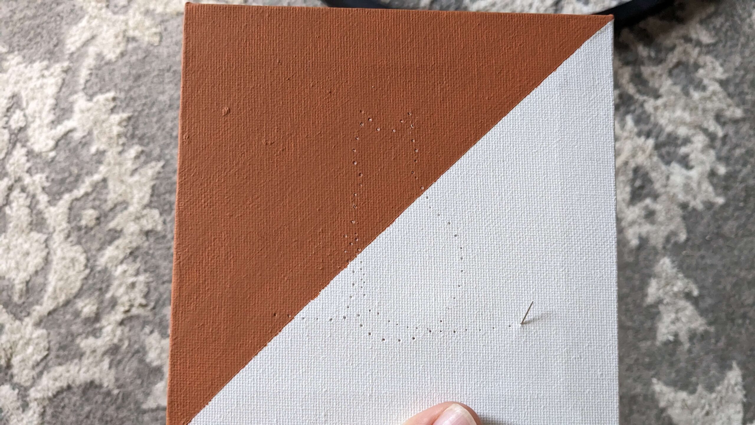 a needle poking through a hole in a square canvas that is diagonally painted half orange half white