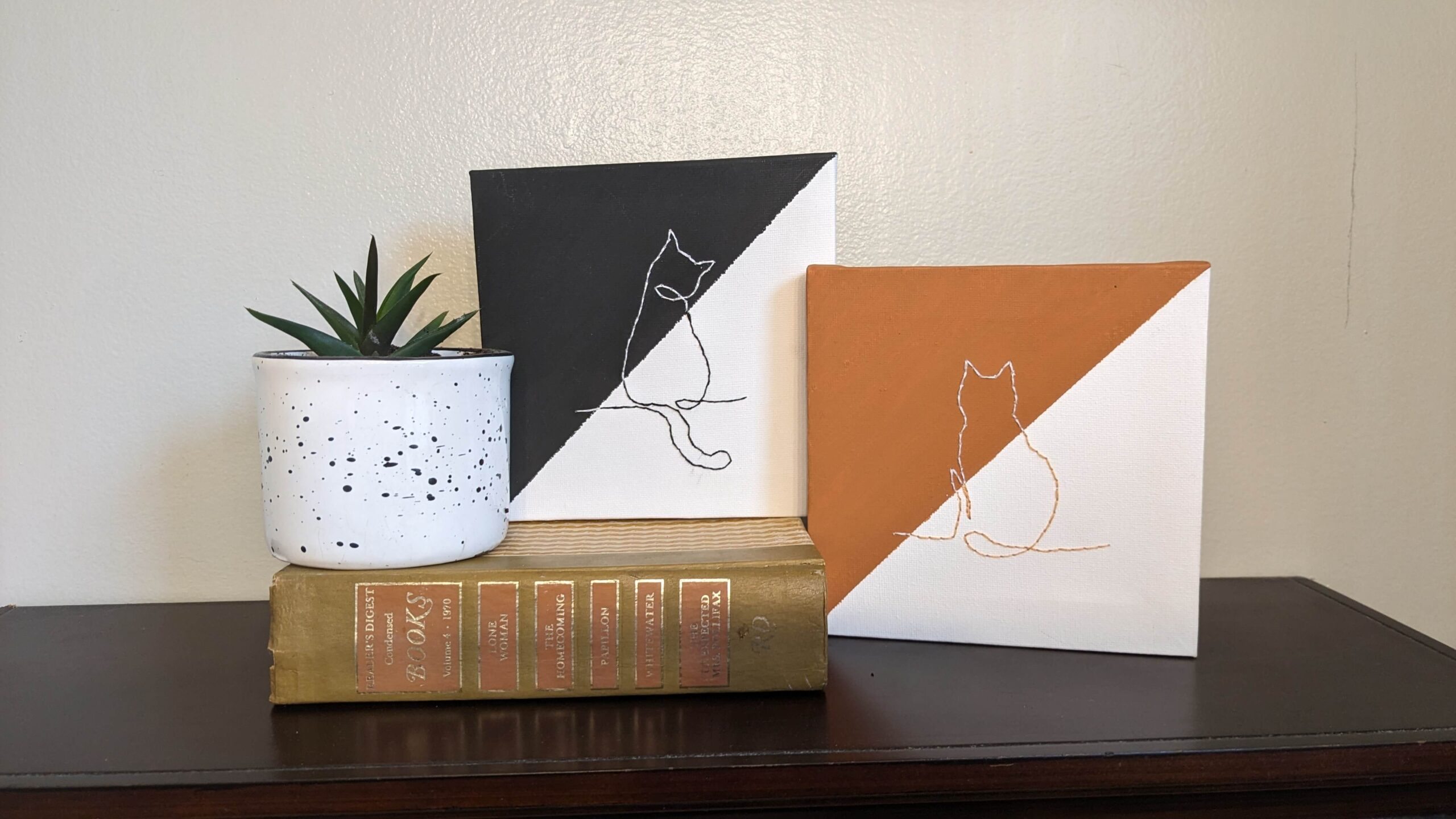 Two pet wall art canvases with cats embroidered on them on a brown shelf with a plant and book