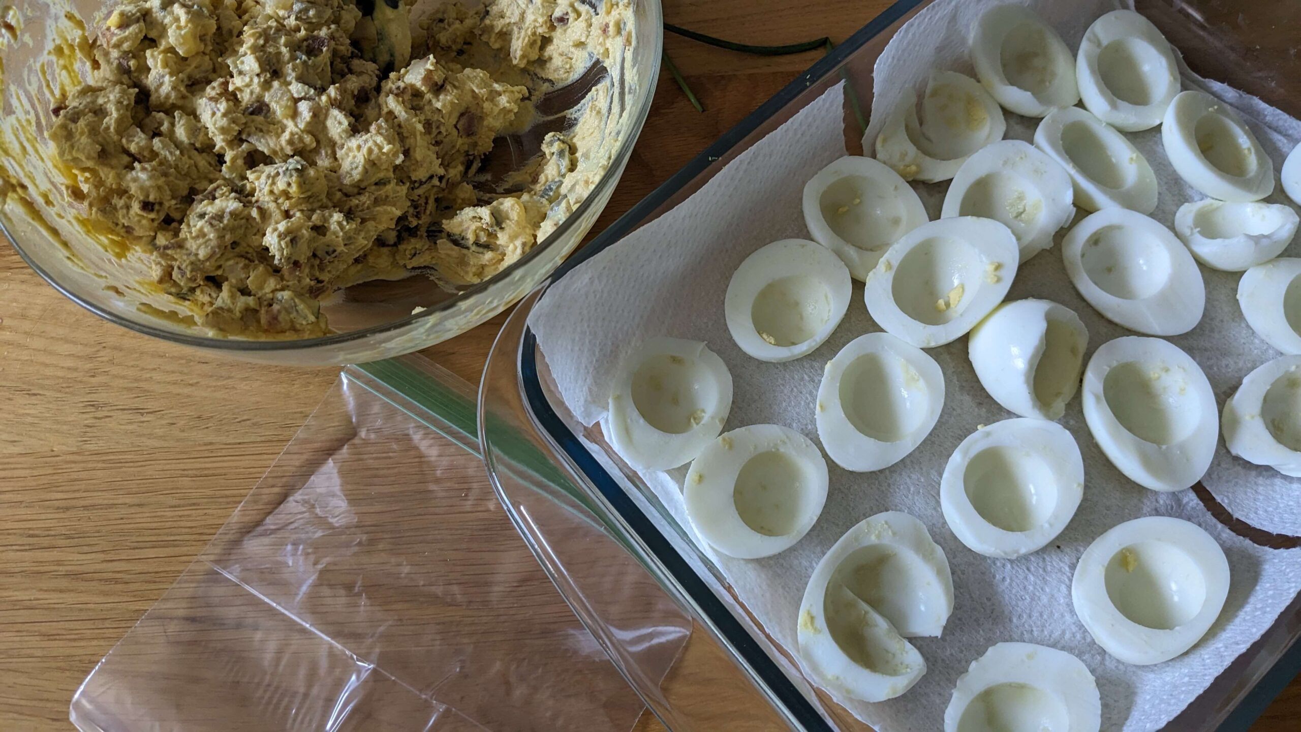 glass bowl of deviled egg mixture next to a tray of hard boiled egg white halves and a ziplock bag
