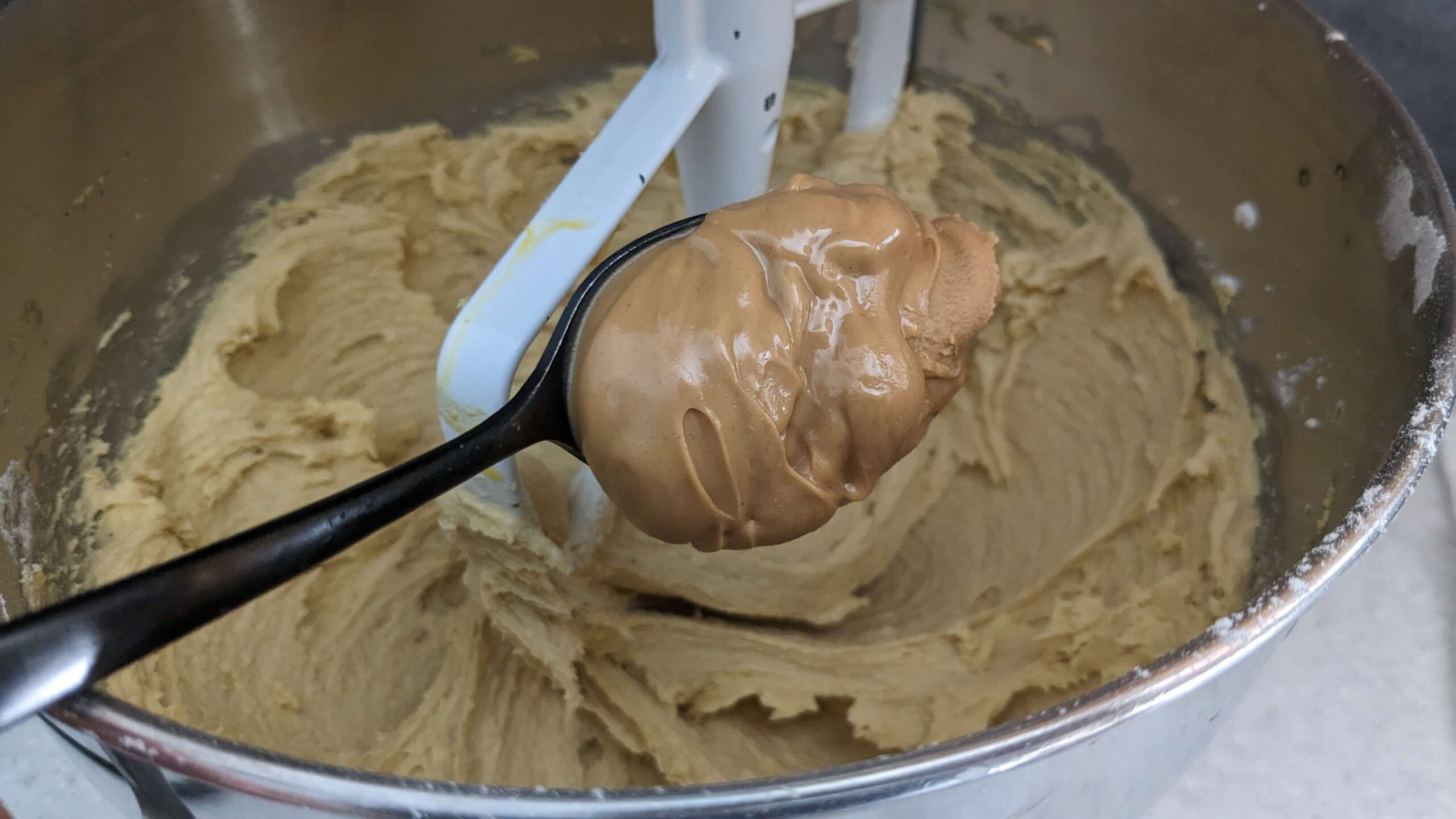 a spoonful of peanut butter being dropped into a kitchen aid mixer bowl of cookie dough
