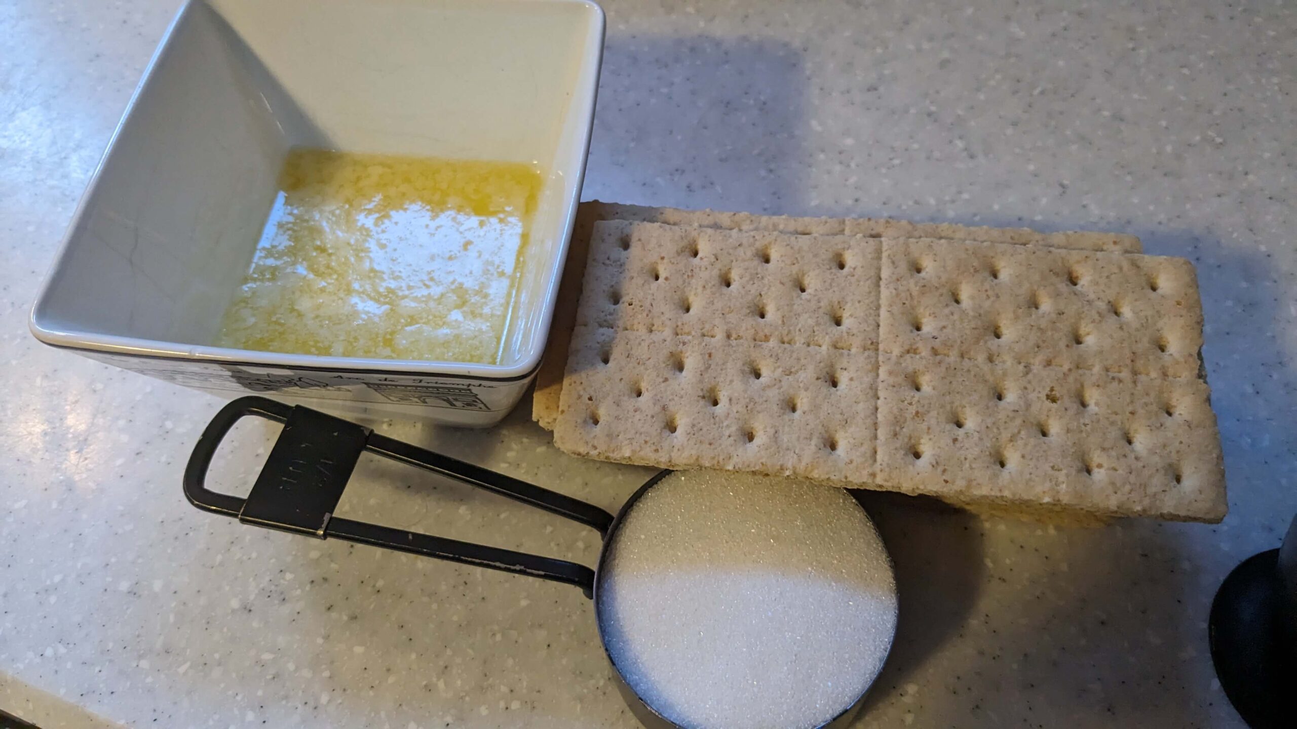bowl of melted butter next to a stack of graham crackers next to a measuring cup of sugar