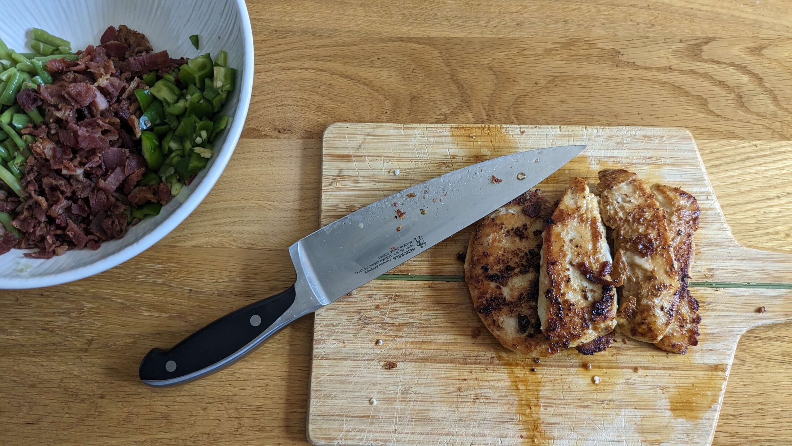 a white bowl filled with chopped green beans, bacon, and green peppers next to a wooden cutting board with four cooked chicken breasts and a chefs knife