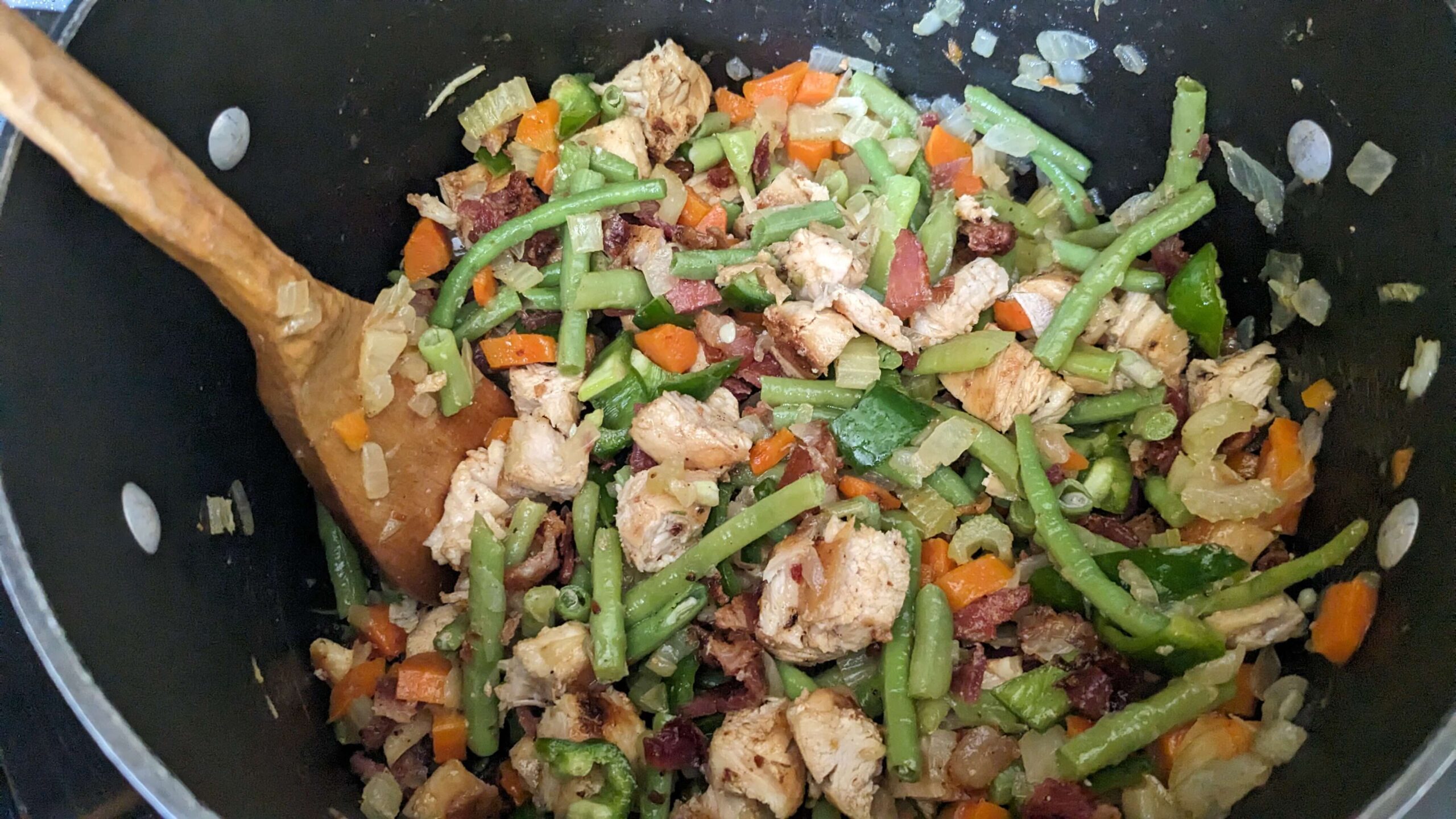 a pot filled with sauteing chopped cooked chicken, diced peppers, diced cooked bacon, and chopped green beans and a wooden spatula