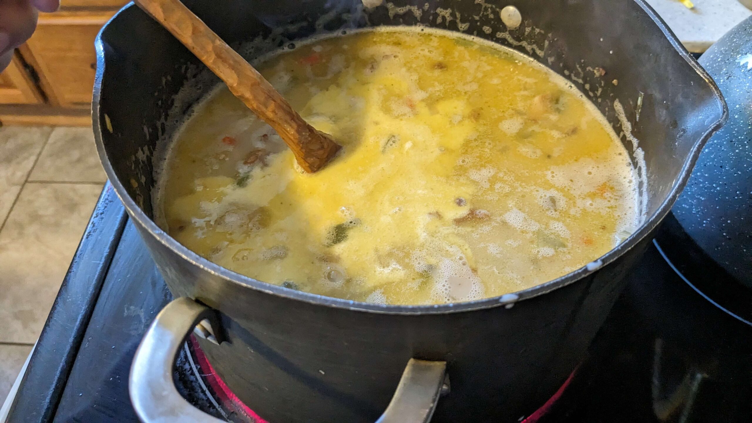 a pot on the stove with a wooden utensil in creamy liquid with diced vegetables near the surface