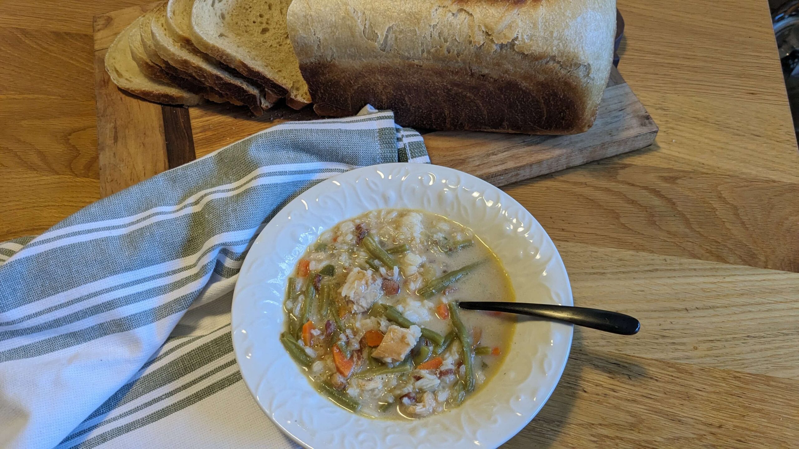 a bowl of creamy chicken, vegetables and rice soup next to a towel and a loaf of sliced sandwich bread on a cutting board 