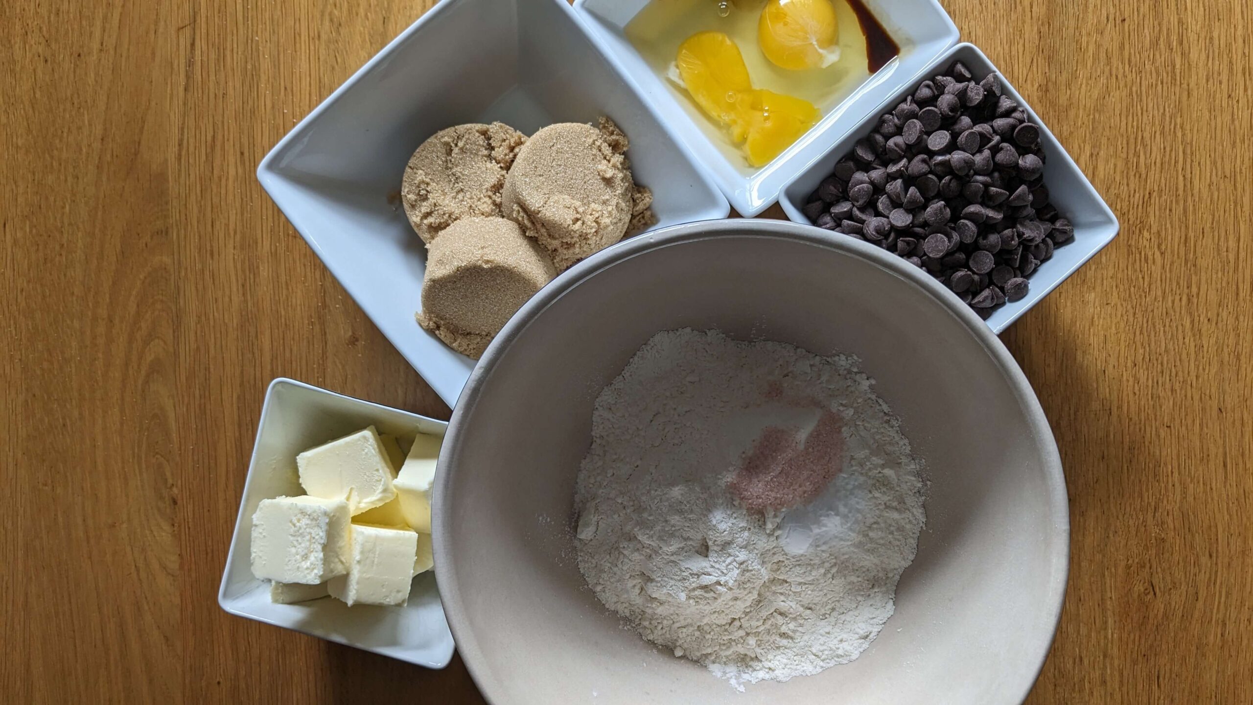 a bowl containing dry baking ingredients, a bowl holding slices of butter, a bowl holding scoops of brown sugar, a bowl holding two egg yokes and whites, and a bowl containing chocolate chips