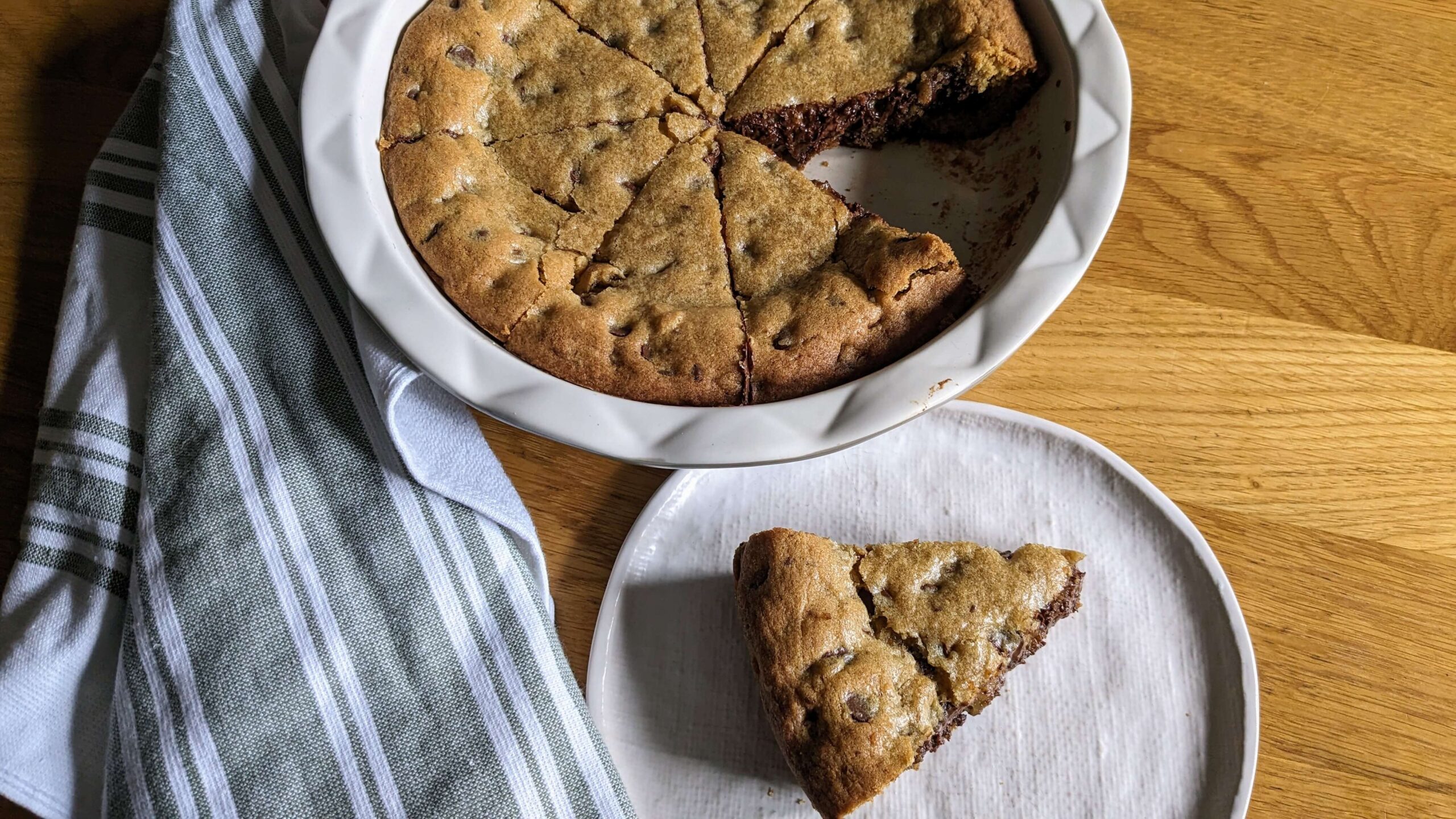a pie plate holding chocolate chip cookie pie with one slice taken out and placed on a white plate next to a green and white towel