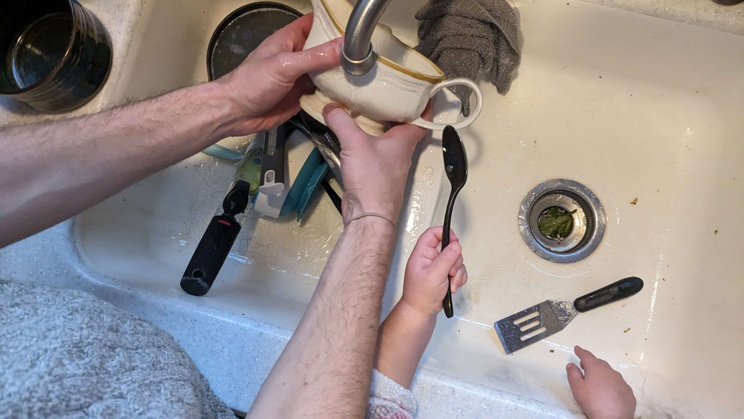 a man washing a dish with a toddler holding a spoon