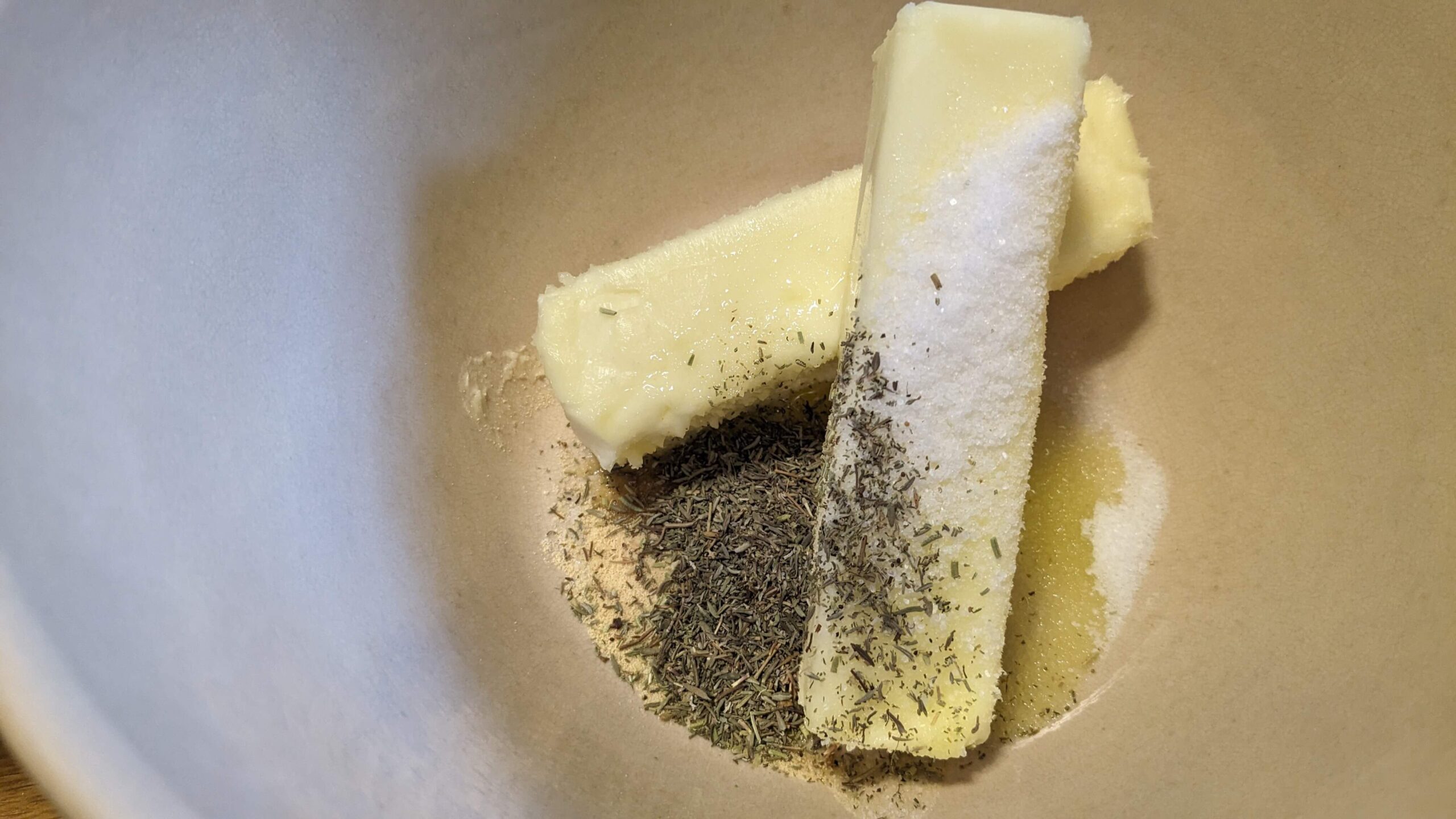 two unwrapped sticks of softened butter in a bowl with herbs