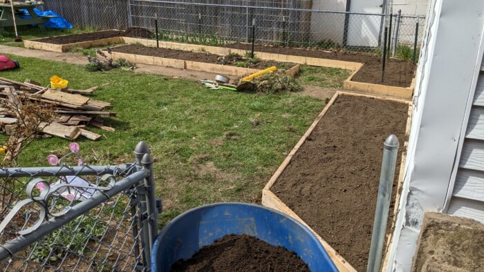 A wheelbarrow is being pushed into a backyard with three empty raised beds.