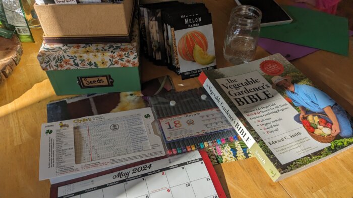 a calendar in front of a box of seeds next to a stack of seed packets pens and the vegetable gardener's bible