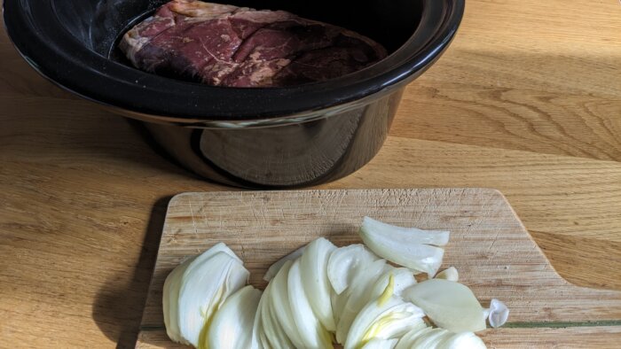 sliced onions on a cutting board next to a crock pot with a roast inside