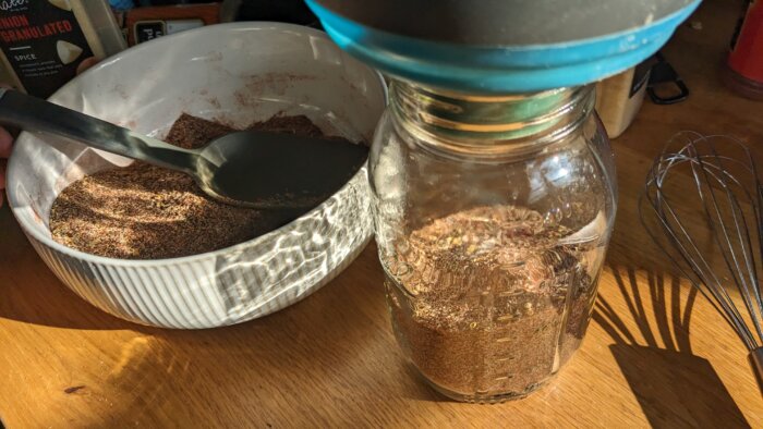 a bowl of spices with a large spoon next to a mason jar half filled with spices and a funnel on top 