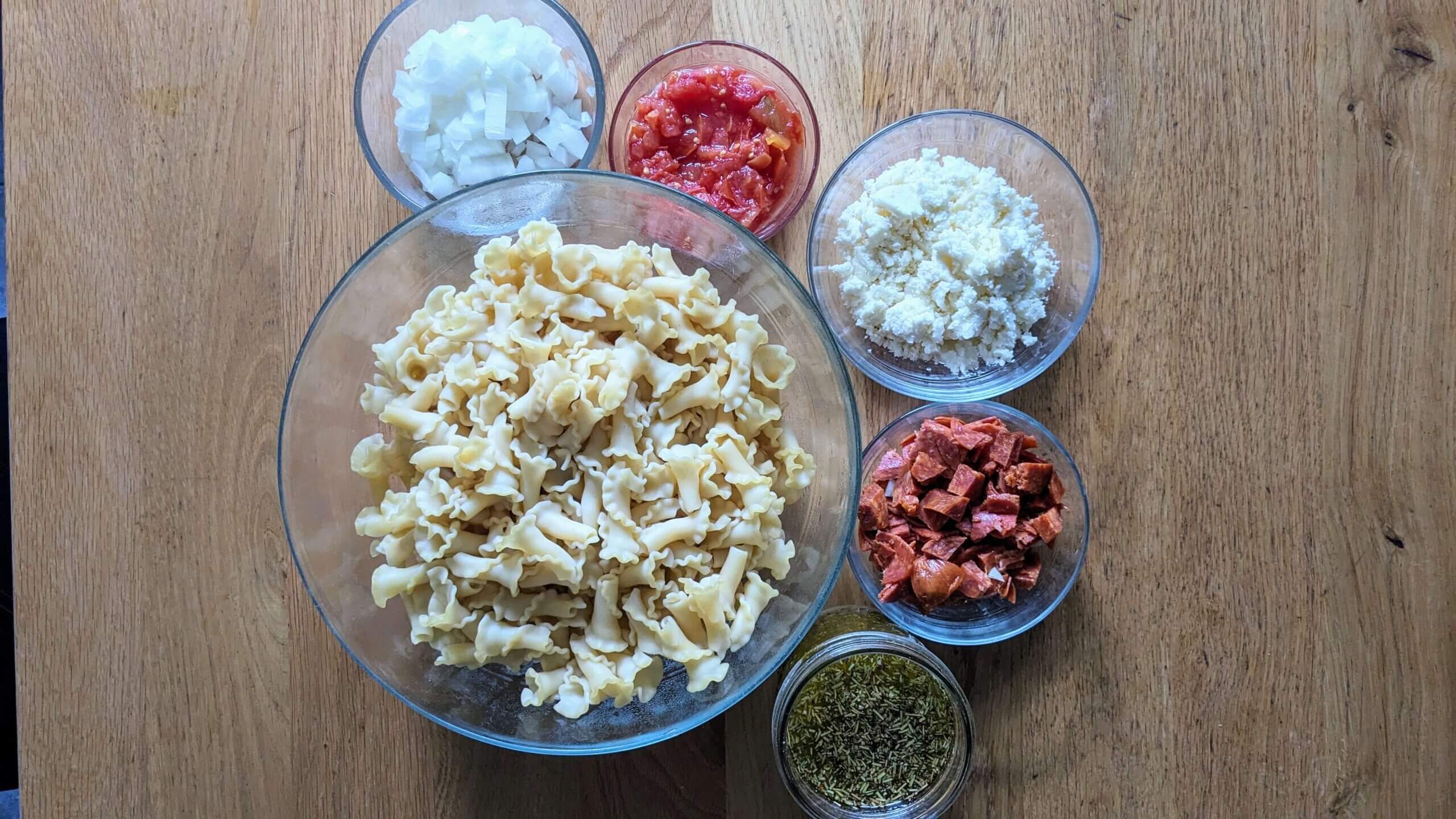 bowl of cooked pasta next to individual bowls of cheese, pepperoni, tomatoes and onins