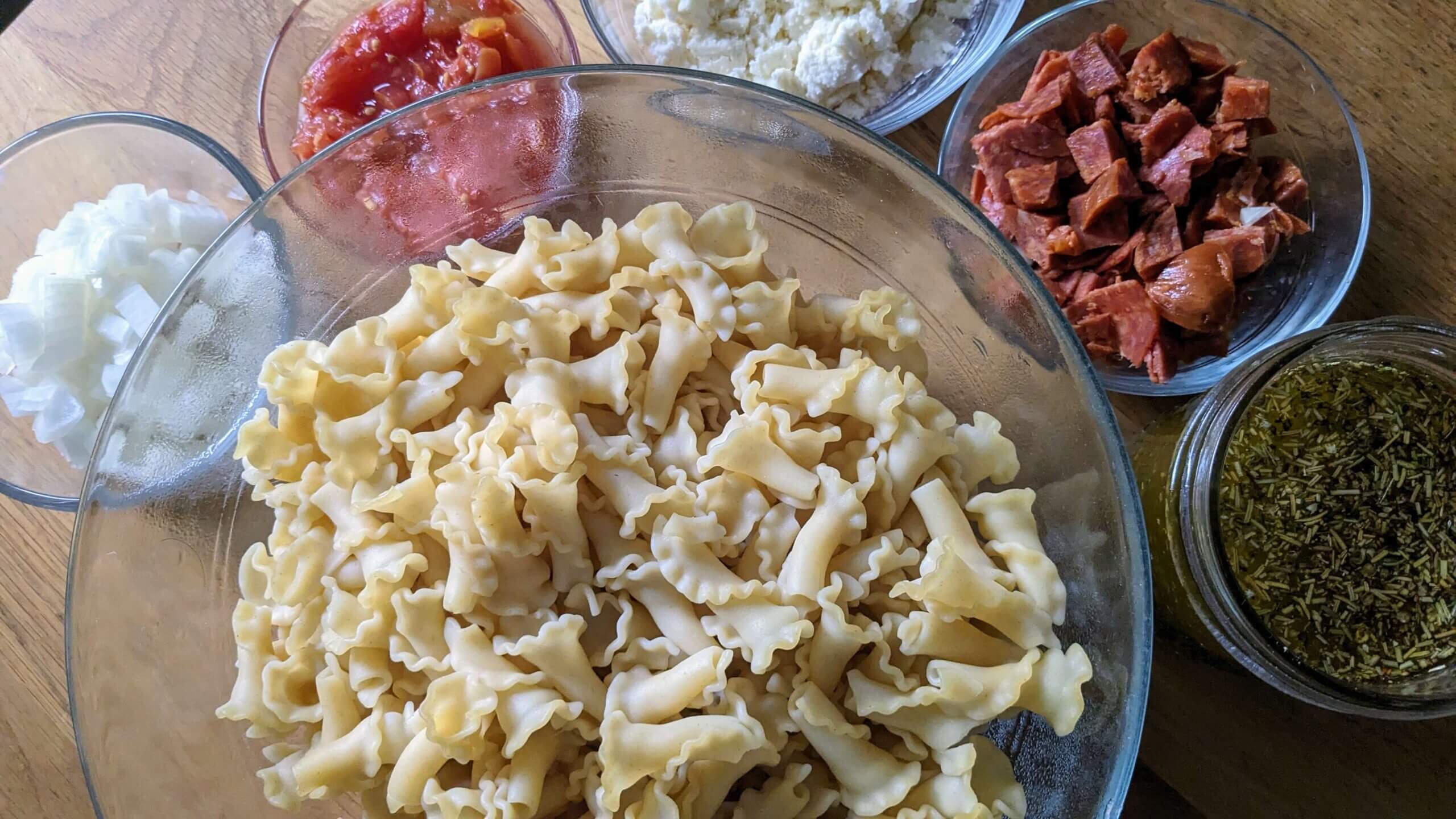 bowl of cooked pasta next to individual bowls of cheese, pepperoni, tomatoes and onins