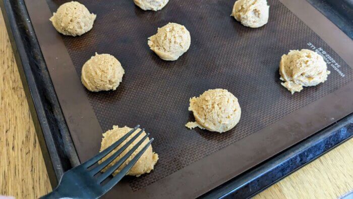 gluten free peanut butter cookie batter in balls on a sil pat on a cookie sheet with a fork pushing into one ball of dough