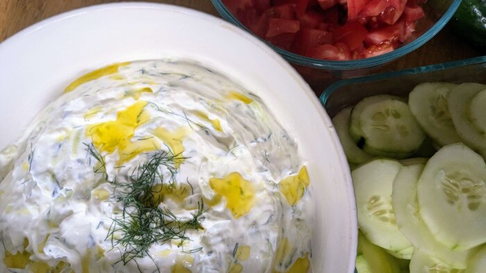 tzatziki sauce topped with olive oil and dill next to bowls of cucumber and tomato