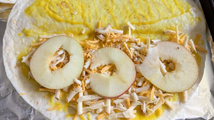 honey mustard on a tortilla wrap topped with shredded cheddar and smoke Gouda cheese and thinly sliced apples