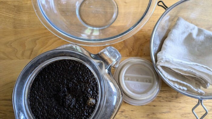 a pitcher of cold brew coffee next to a fine mesh strainer, cheesecloth, and empty glass bowl