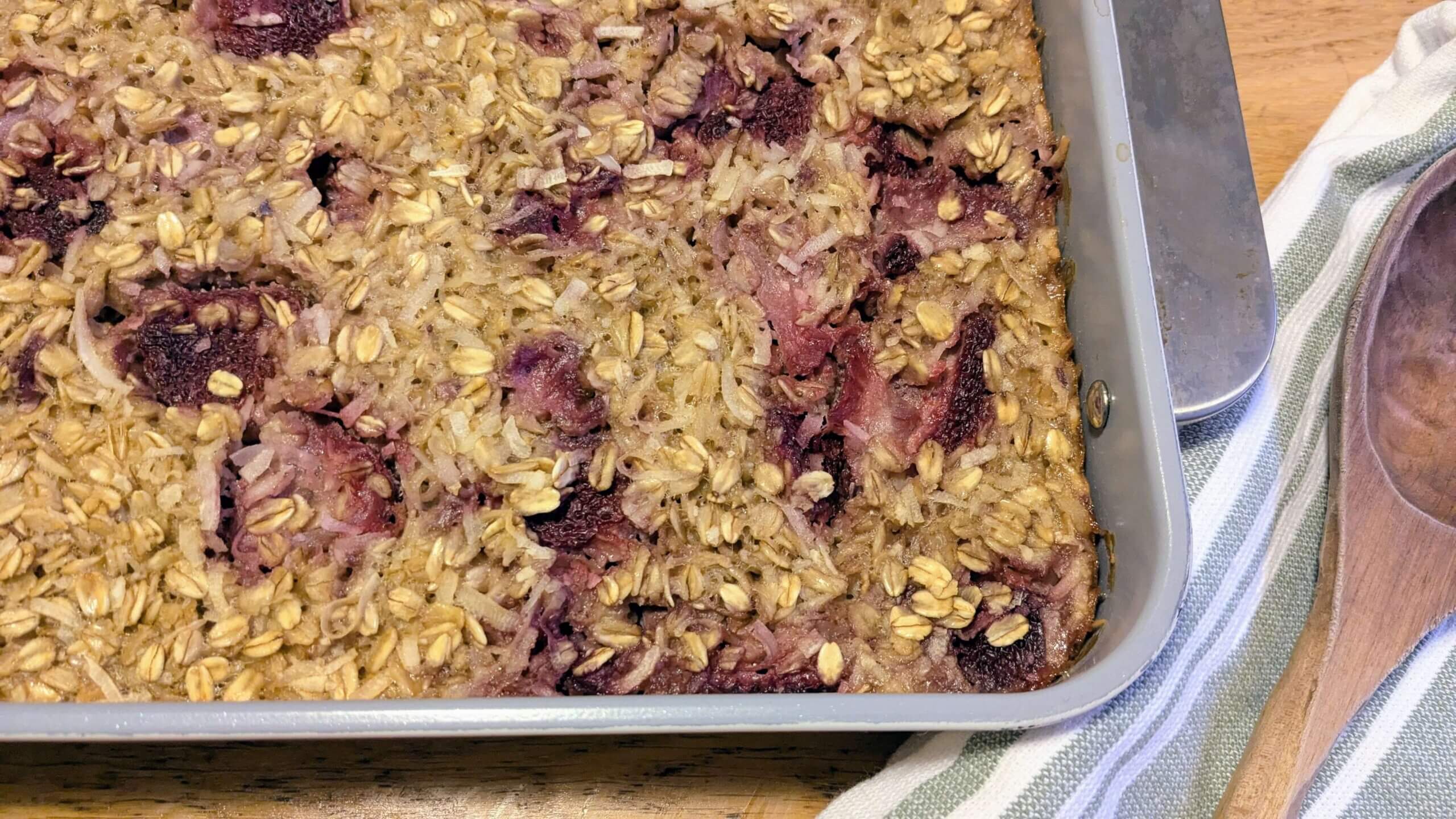 Strawberry coconut oatmeal bake in a 9x13 pan next to a wooden spoon on a towel