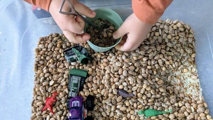 toddler boy playing in a sensory bin with a cup, spoons, dinosaurs and toy cars