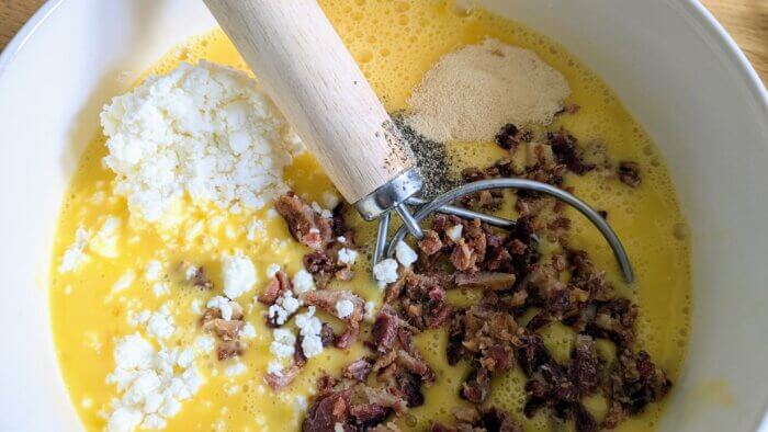 uncooked scrambled eggs in a white bowl with a Danish dough hook, feta cheese, crumbled bacon, garlic powder, and black pepper