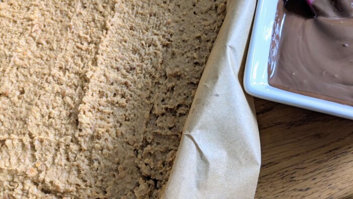 peanut butter and pretzel mixture pressed into a parchment paper lined 9x13 pan next to a bowl of melted chocolate