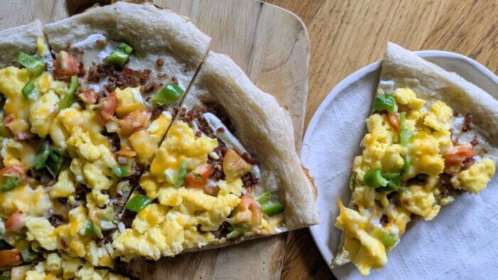 a slice of breakfast pizza on a plate next to a cutting board of breakfast pizza