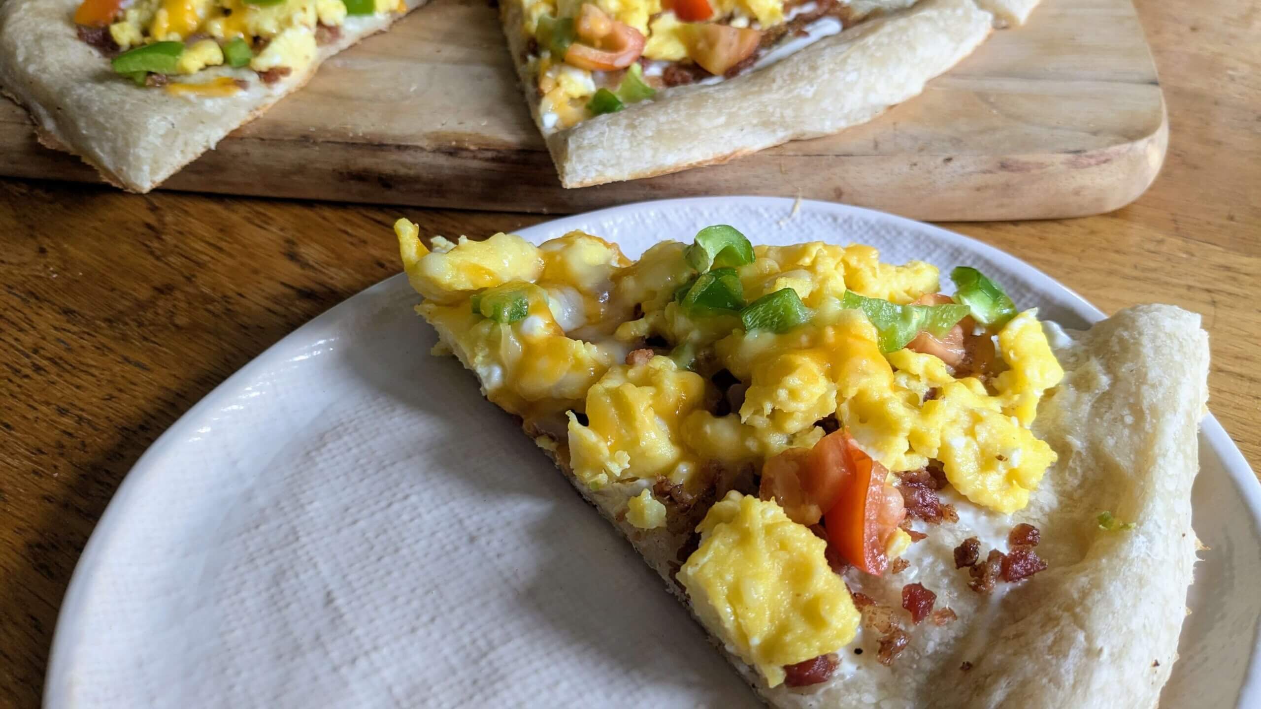 a slice of breakfast pizza on a plate next to a cutting board of breakfast pizza