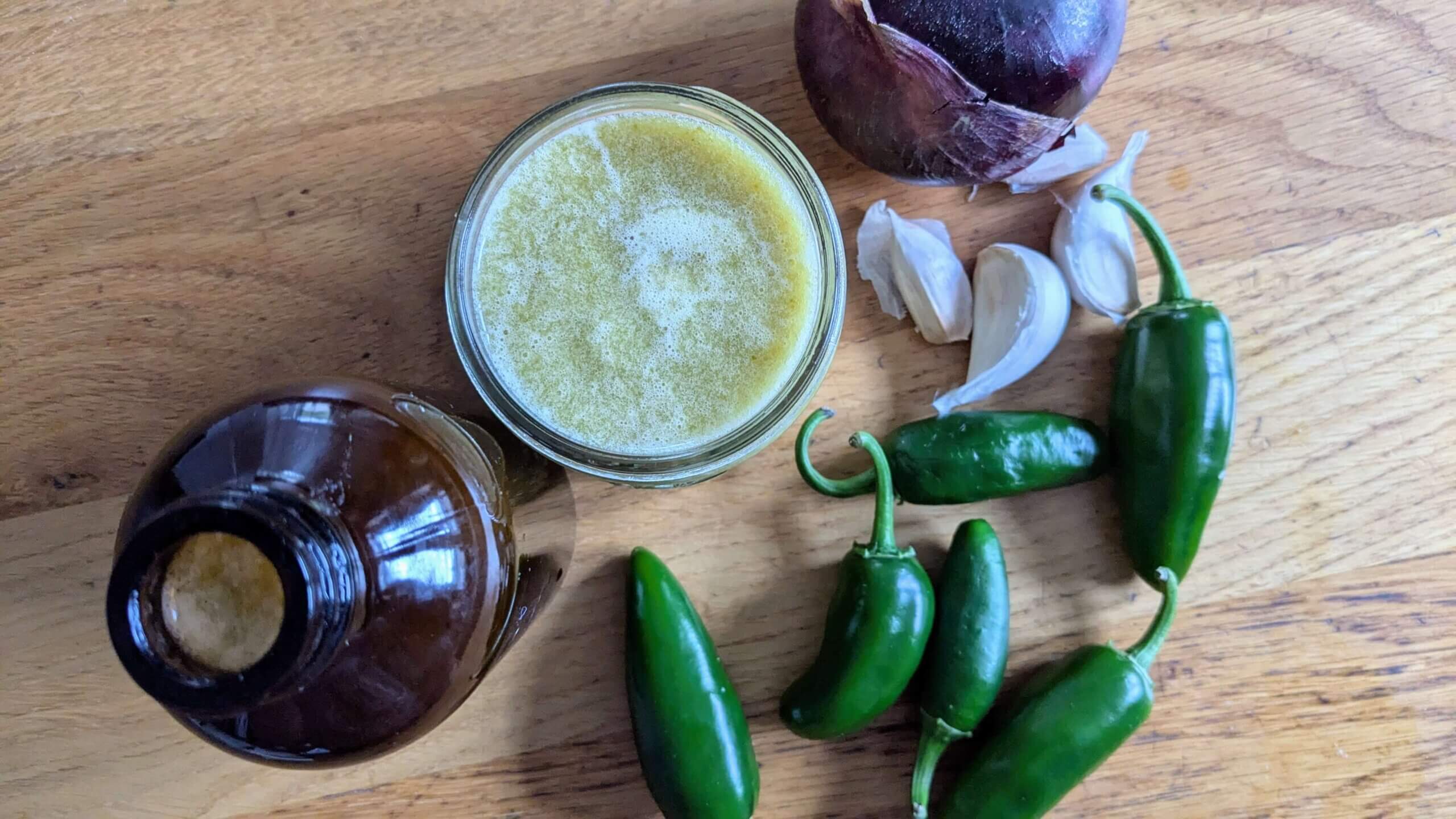 jalapeno Hot sauce in a mason jar surrounded by jalapenos, garlic, and a red onion