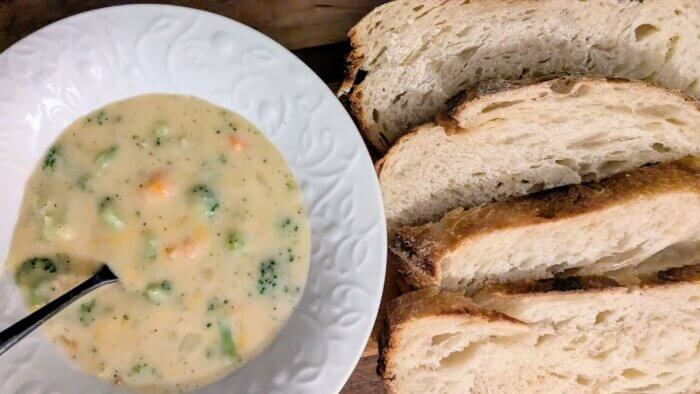 a white bowl of broccoli cheddar soup next to sliced sourdough bread