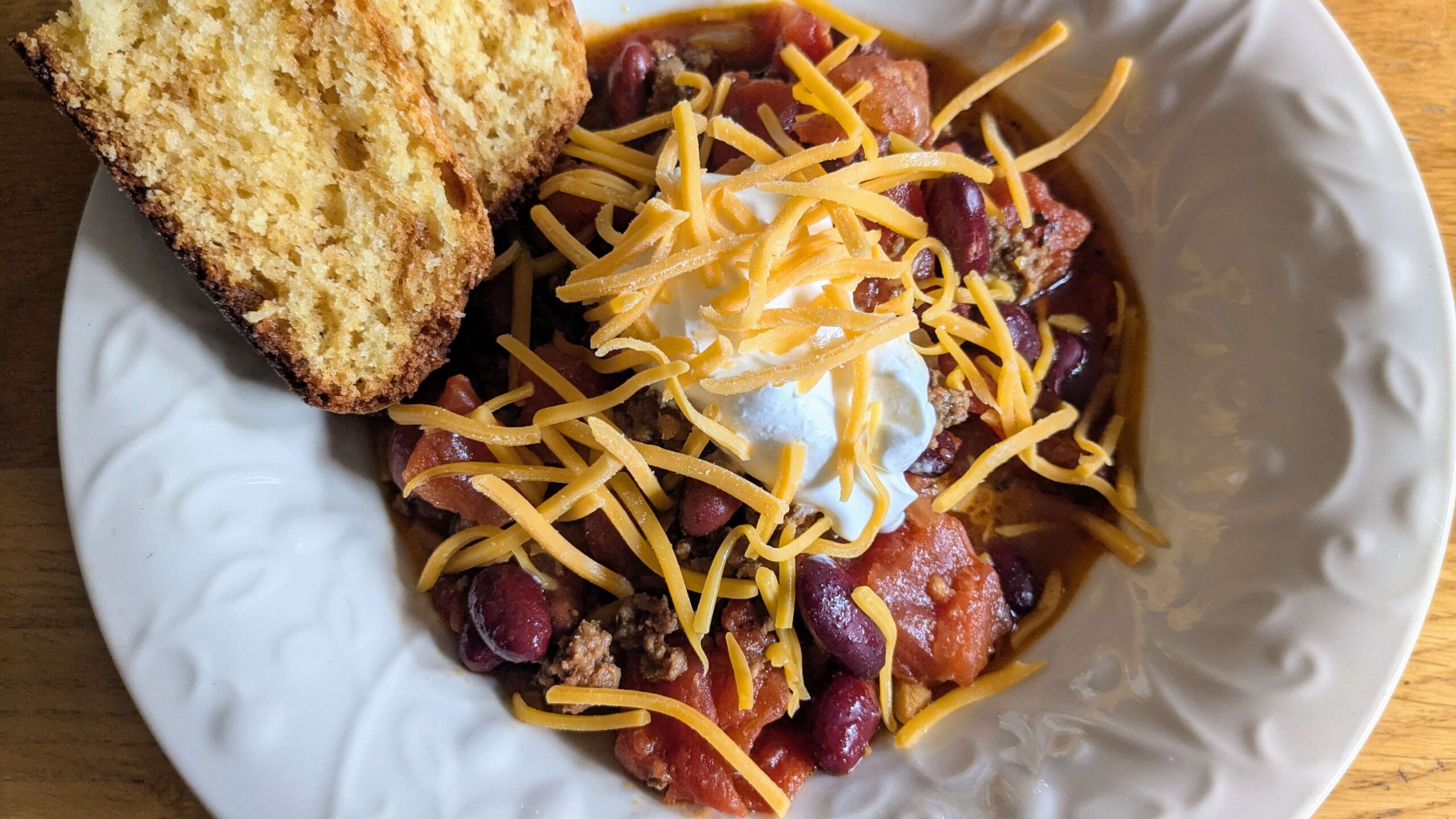 a bowl of crockpot chili topped with sour cream, cheese, and a side of cornbread